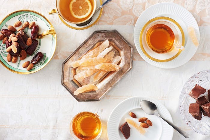 Pomelo candies on antique metal plate alongside orange tea and dishes of almonds, chocolate and dates, atop pale floral tablecloth.