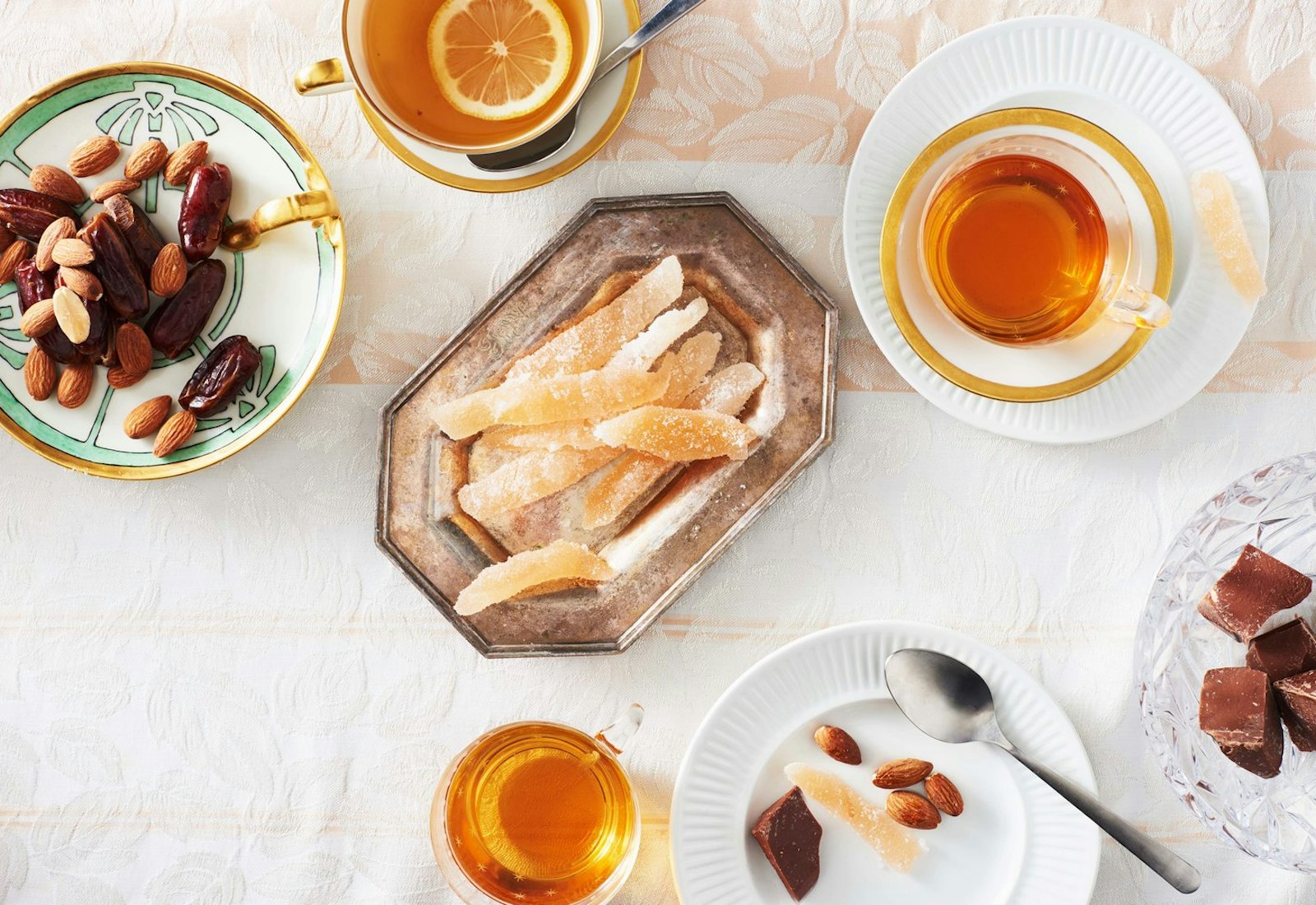 Pomelo candies on antique metal plate alongside orange tea and dishes of almonds, chocolate and dates, atop pale floral tablecloth.
