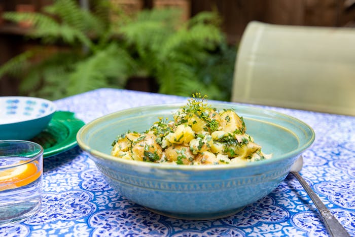 Potato salad with fresh herbs in blue ceramic bowl atop blue patterned tablecloth, outdoor scene.
