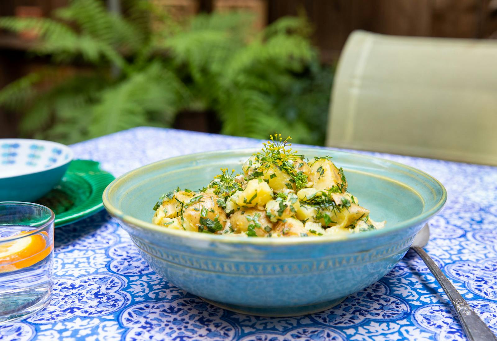 Potato salad with fresh herbs in blue ceramic bowl atop blue patterned tablecloth, outdoor scene.