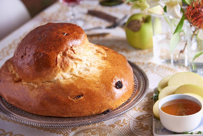 Round raisin challah on dining table alongside sliced green apples, a bowl of honey and fresh flowers.