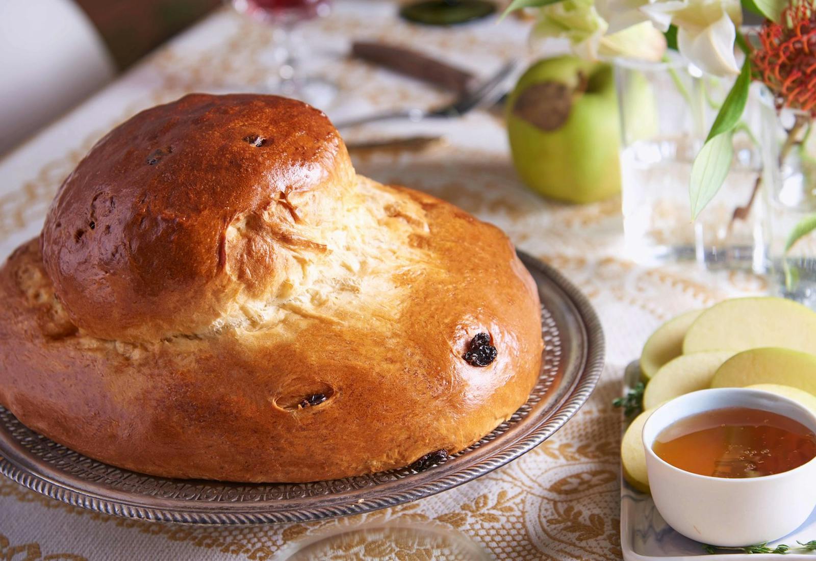 Round raisin challah on dining table alongside sliced green apples, a bowl of honey and fresh flowers.