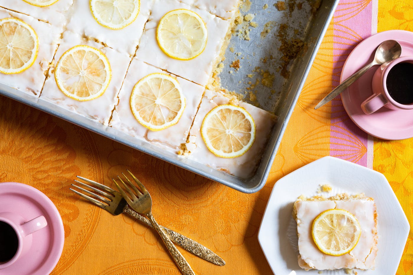 Lemon velvet sheet cake sliced into portions in large baking dish alongside mugs of coffee, atop bright orange and pink tablecloth.