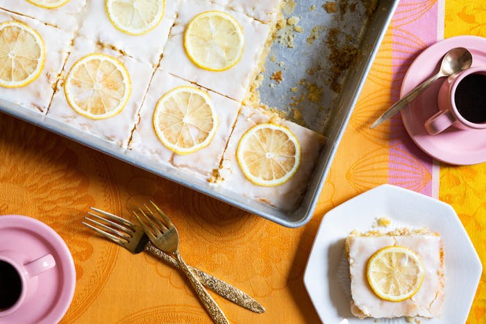 Lemon velvet sheet cake sliced into portions in large baking dish alongside mugs of coffee, atop bright orange and pink tablecloth.