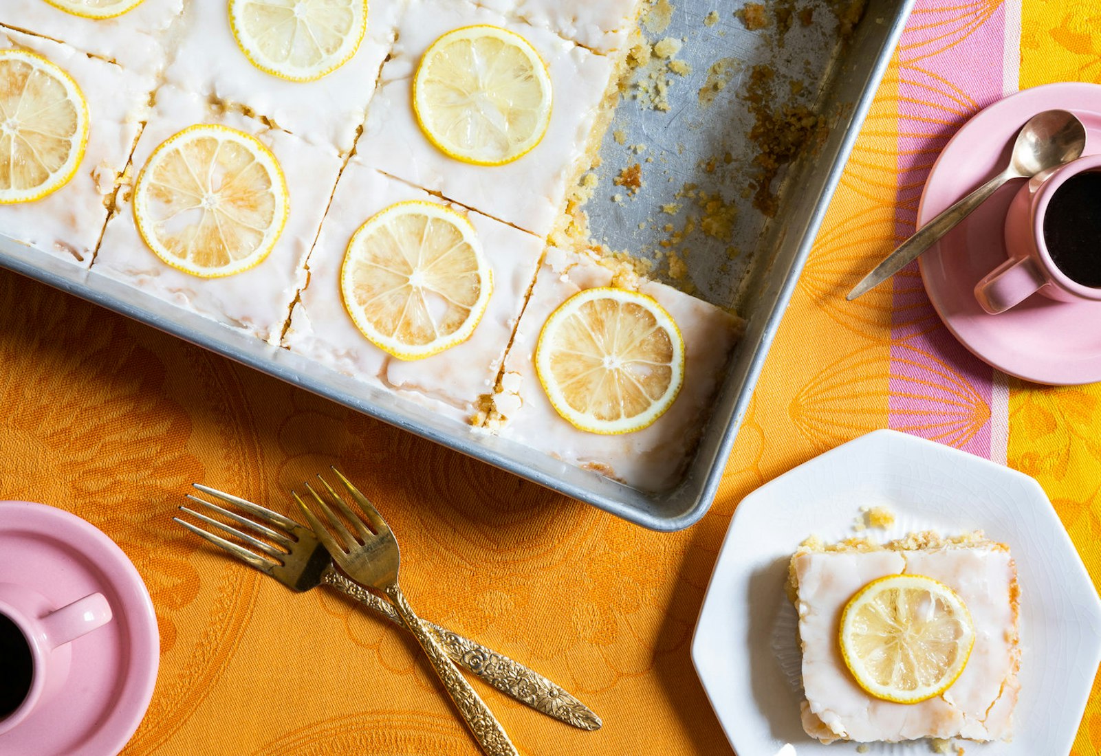 Lemon velvet sheet cake sliced into portions in large baking dish alongside mugs of coffee, atop bright orange and pink tablecloth.