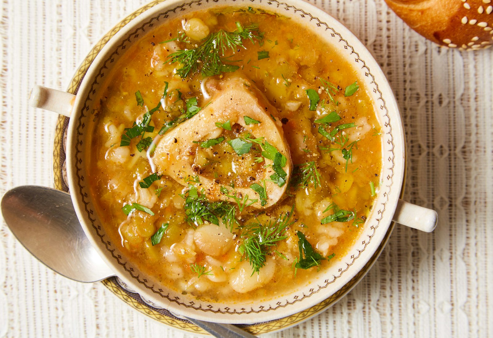 Split pea soup with marrow bone in serving bowl with handles alongside seeded challah atop white tablecloth.
