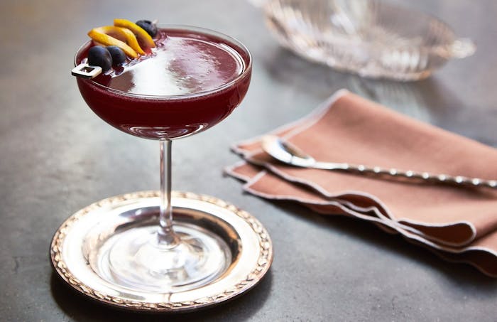 Blueberry cocktail garnished with blueberries and lemon slices, atop black surface.
