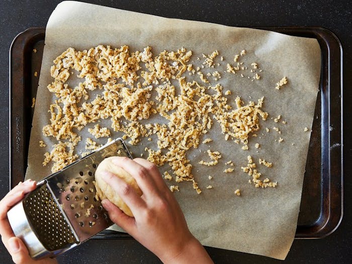 Sheet pan with parchment paper and fresh barley, person grating dough on cheese grater. 