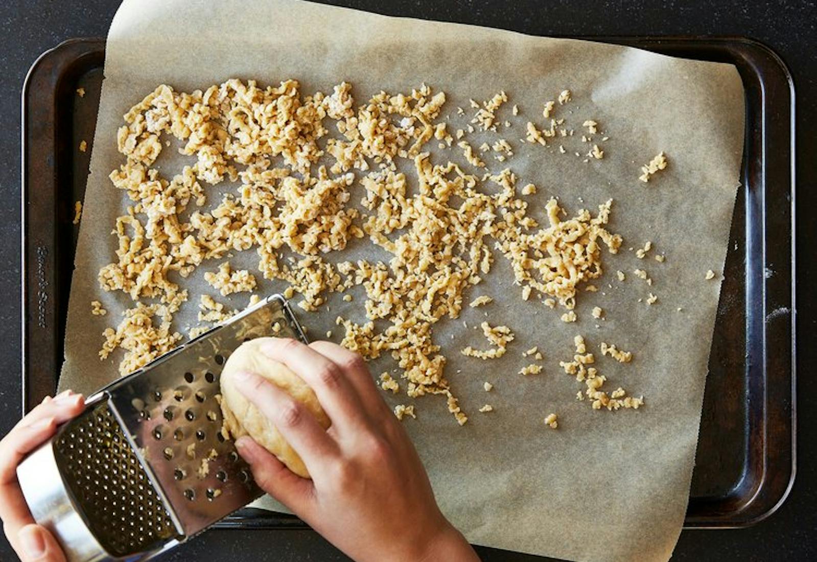 Sheet pan with parchment paper and fresh barley, person grating dough on cheese grater. 