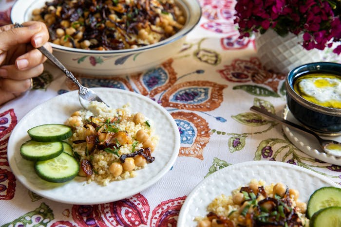 Bulgur with fried onions and sliced cucumber alongside bowl of cucumber mint yogurt, atop watercolor tablecloth.