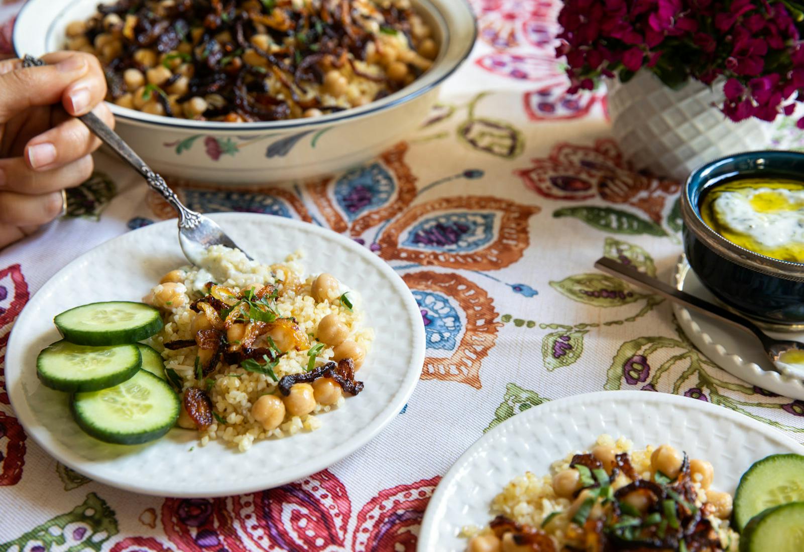 Bulgur with fried onions and sliced cucumber alongside bowl of cucumber mint yogurt, atop watercolor tablecloth.