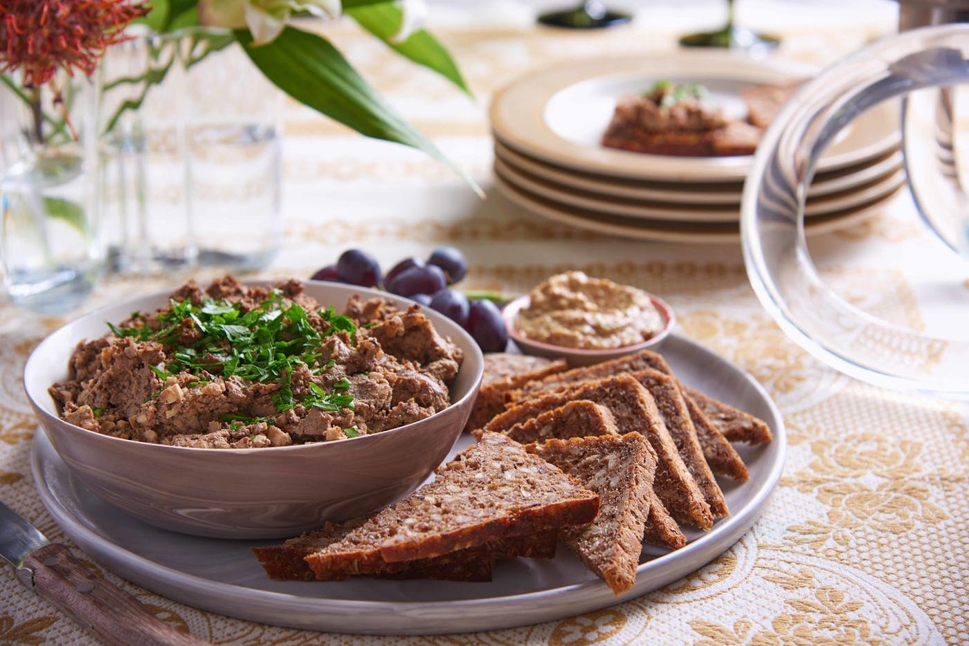 Chopped liver with chopped parsley alongside sliced seeded bread, grainy mustard and grapes, with fresh flower vase atop orange patterned tablecloth.