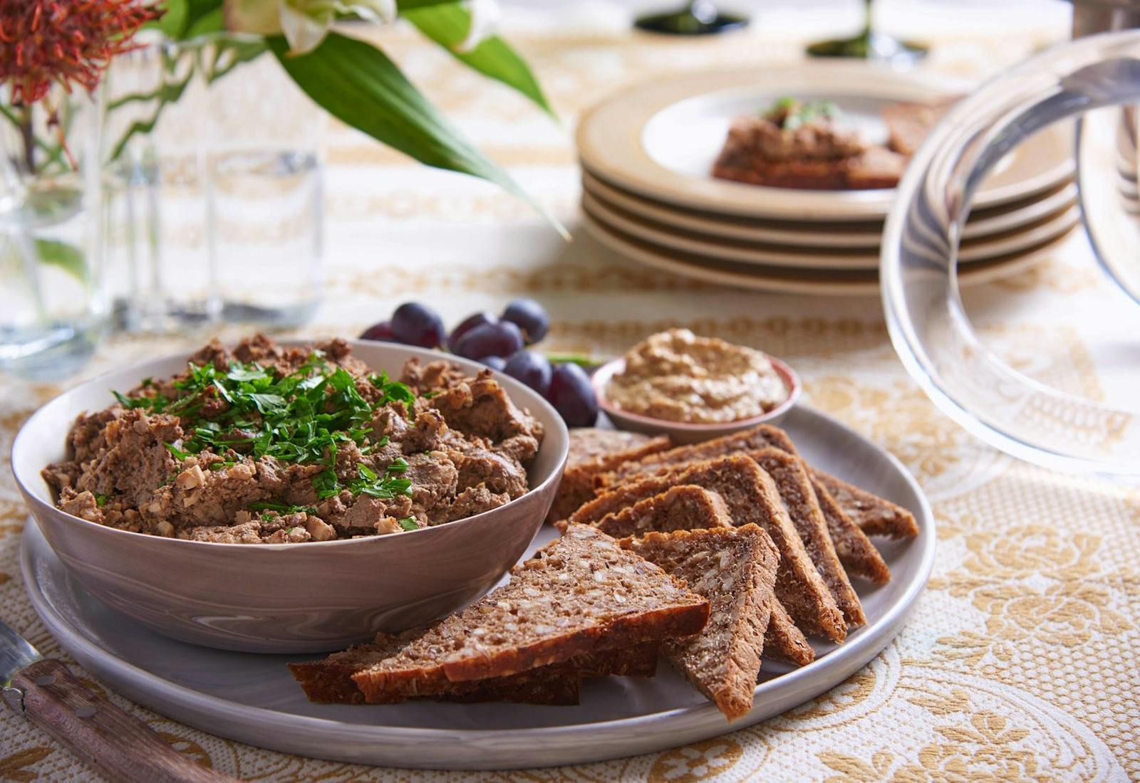 Chopped liver with chopped parsley alongside sliced seeded bread, grainy mustard and grapes, with fresh flower vase atop orange patterned tablecloth.