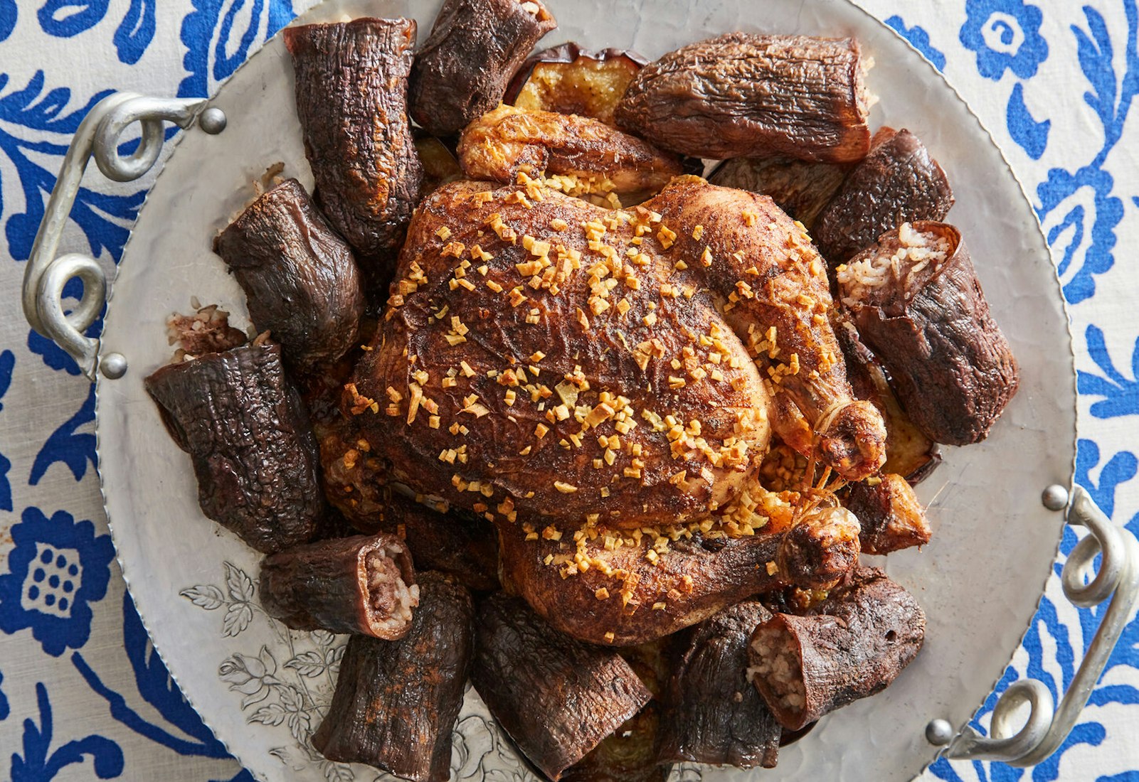 Roasted chicken surrounded by stuffed eggplants on large tray atop blue floral tablecloth.