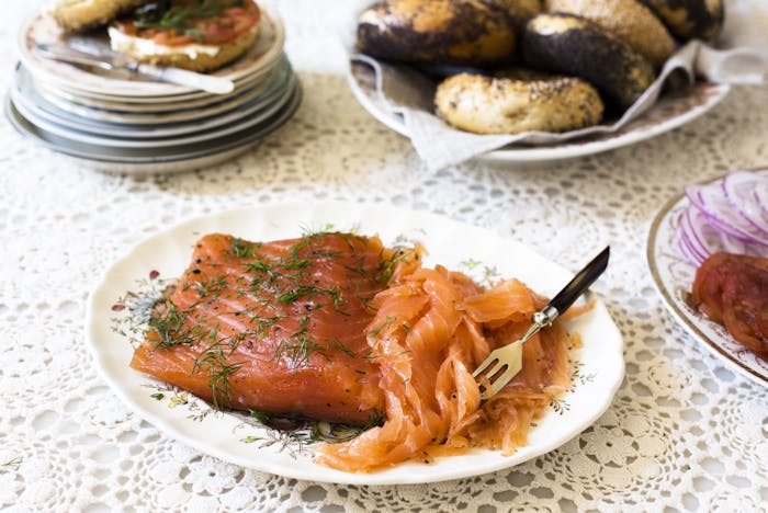 Plate with cured filet of salmon and sliced gravlax garnished with dill alongside plates of poppyseed bagels, tomatoes and sliced red onions atop woven white tablecloth.