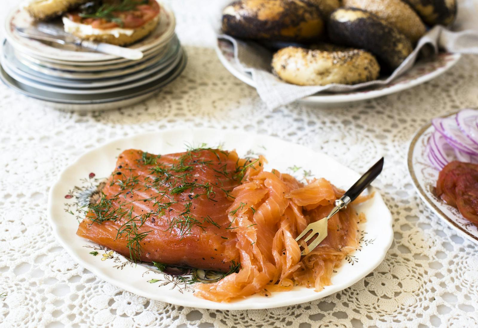 Plate with cured filet of salmon and sliced gravlax garnished with dill alongside plates of poppyseed bagels, tomatoes and sliced red onions atop woven white tablecloth.
