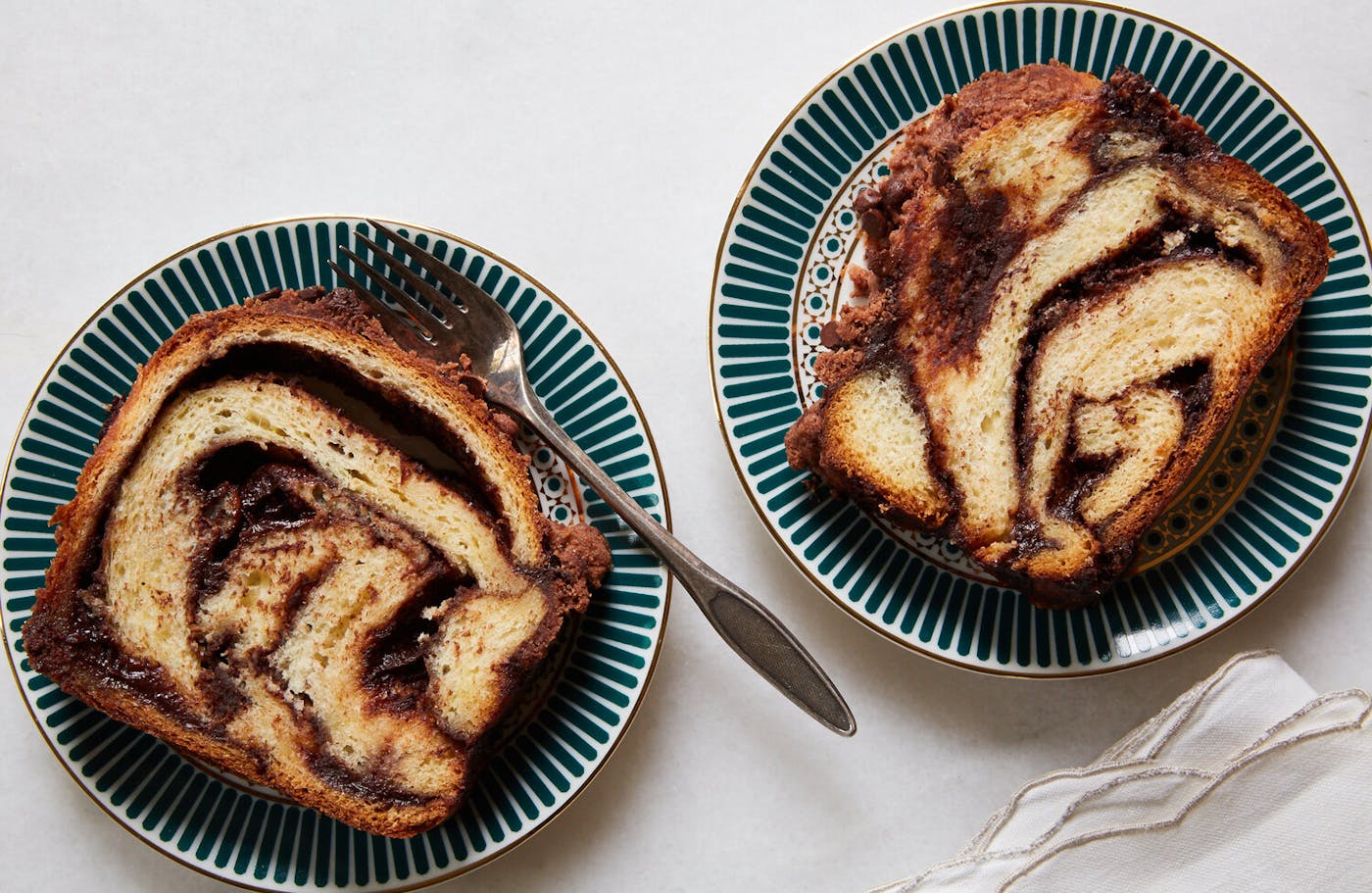 Two slices of chocolate babka on blue and white print plates atop white surface.