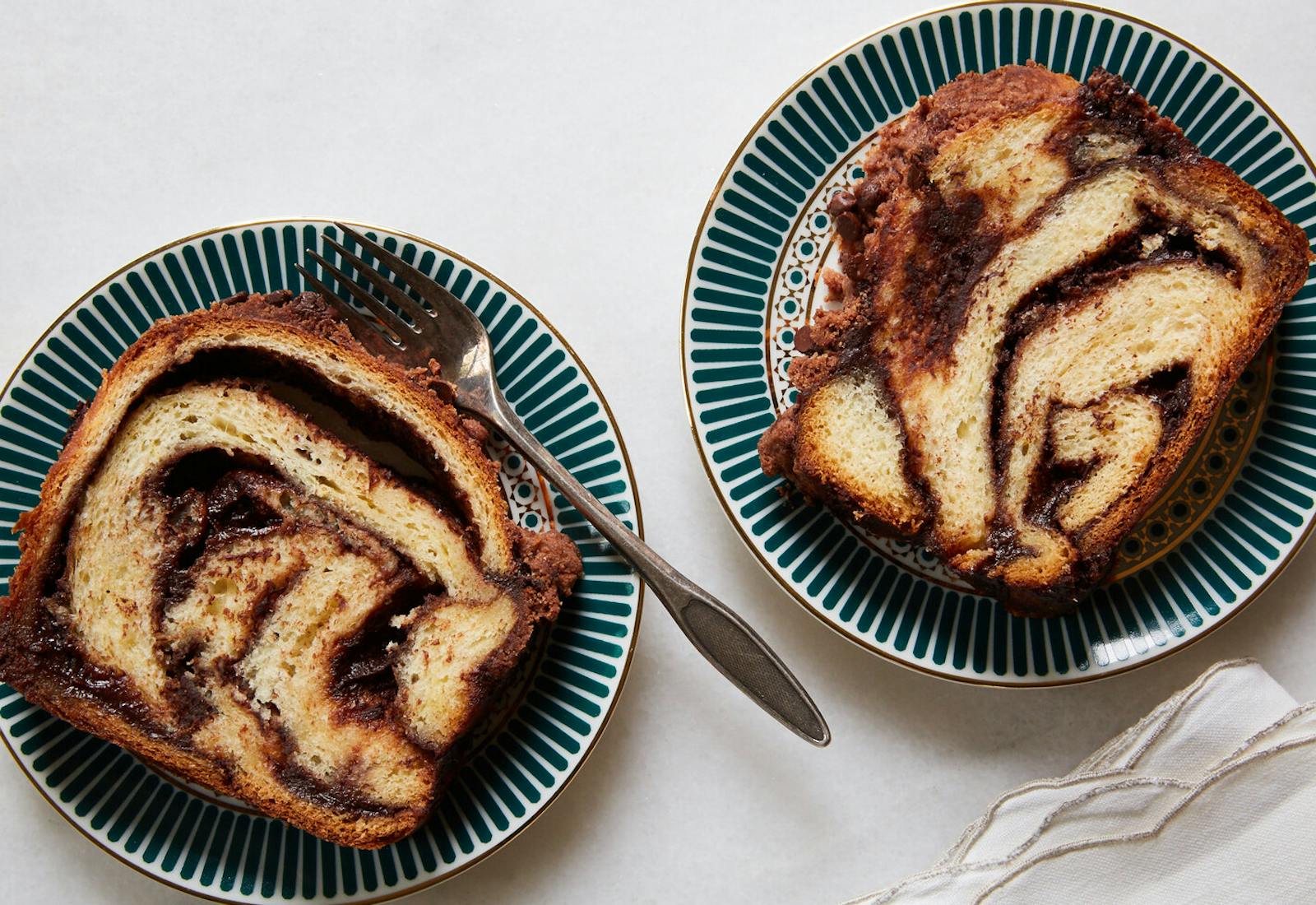 Two slices of chocolate babka on blue and white print plates atop white surface.