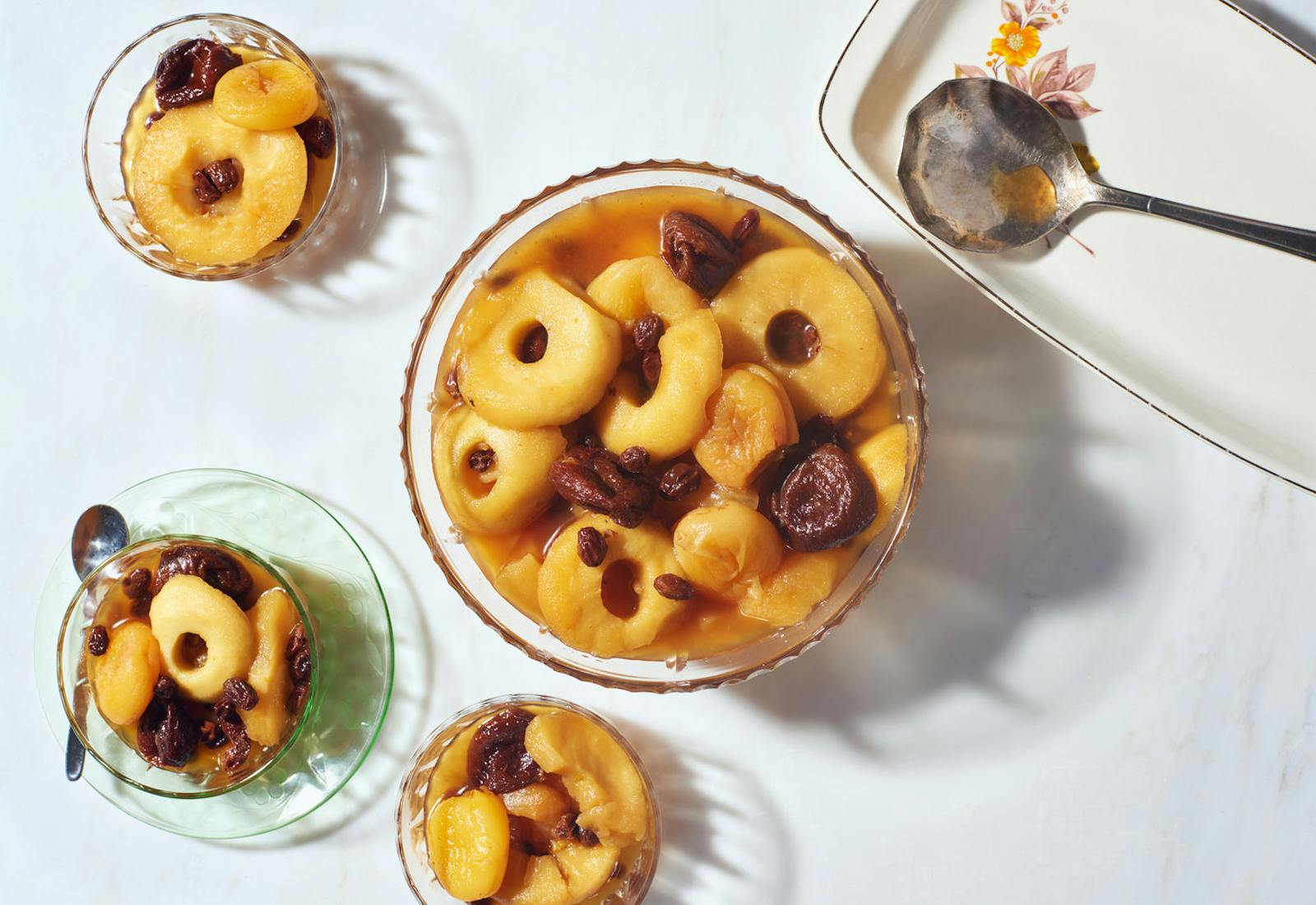 Large and small glass bowls with dried fruit compote atop white surface.