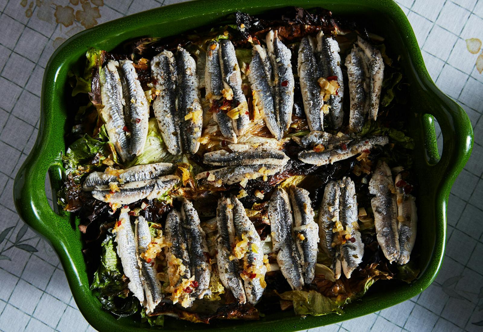 Baked anchovies on a bed of escarole garnished with red chili flakes in green casserole dish, atop grid-patterned tablecloth.