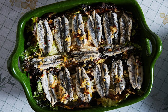 Baked anchovies on a bed of escarole garnished with red chili flakes in green casserole dish, atop grid-patterned tablecloth.