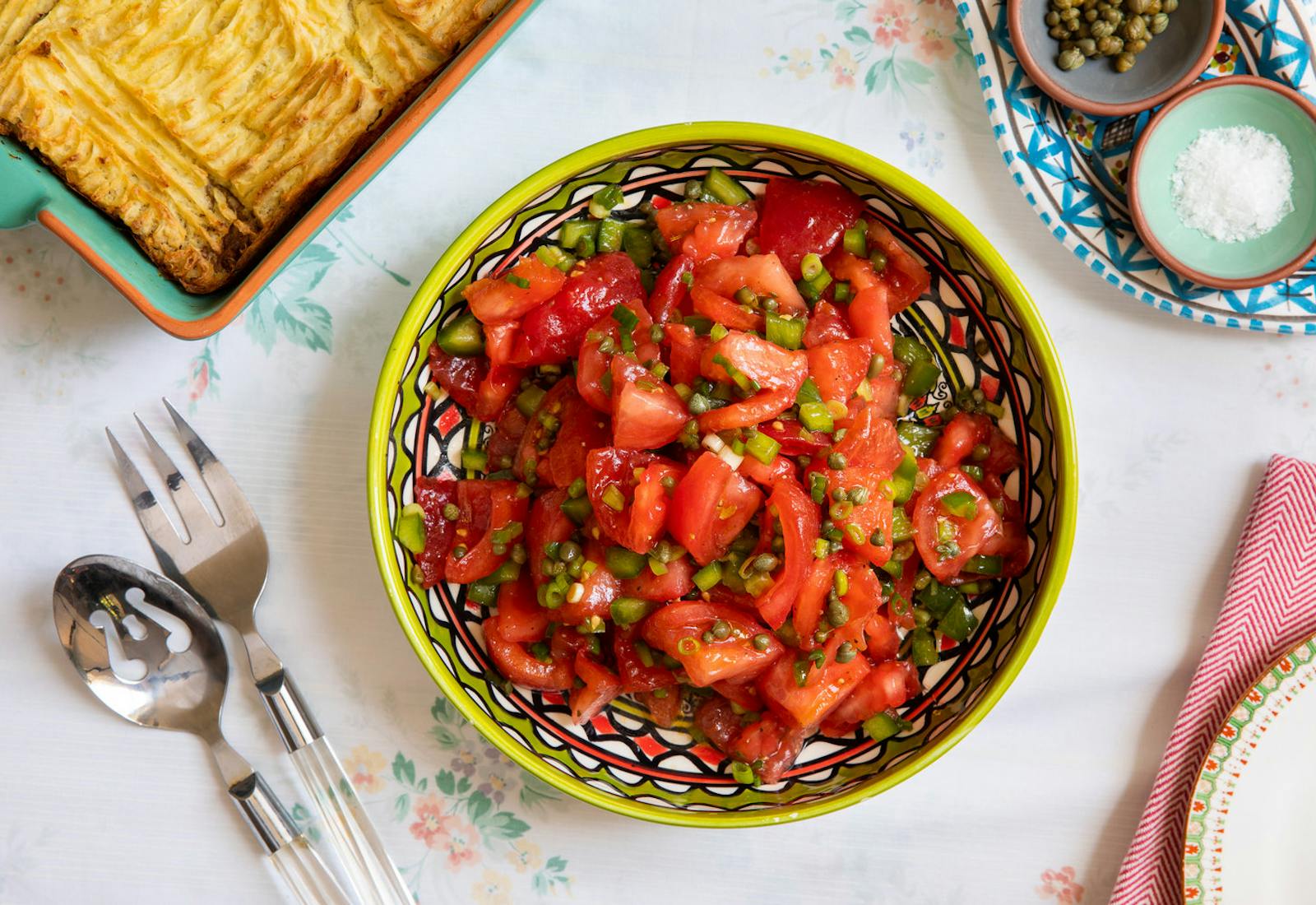 Tomato salad with capers in green and red patterned bowl alongside dishes with coarse salt and capers, atop white floral tablecloth.