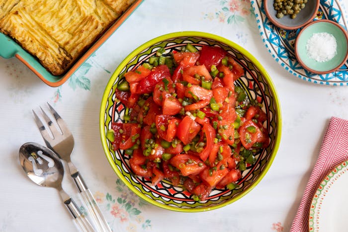 Tomato salad with capers in green and red patterned bowl alongside dishes with coarse salt and capers, atop white floral tablecloth.