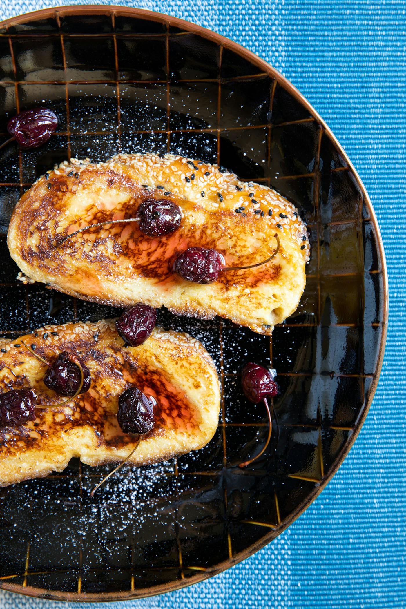 Honey challah with macerated cherries, syrup and powdered sugar on black plate atop blue tablecloth.