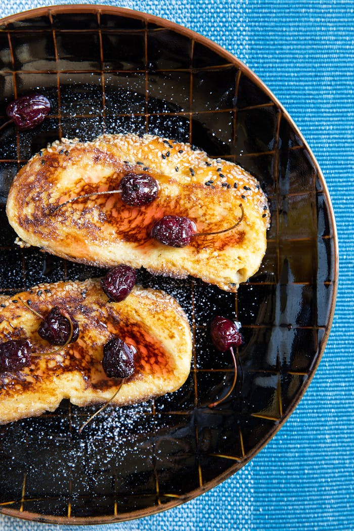 Honey challah with macerated cherries, syrup and powdered sugar on black plate atop blue tablecloth.