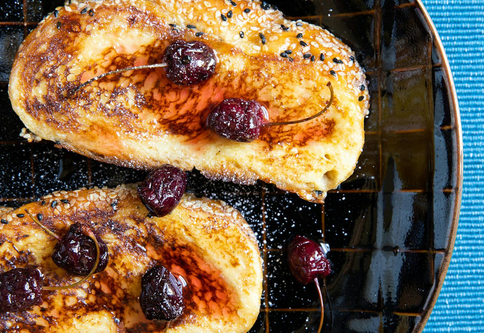 Honey challah with macerated cherries, syrup and powdered sugar on black plate atop blue tablecloth.