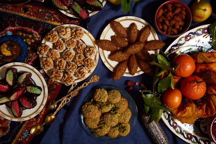 Petits gateaux on blue plate surrounded by various cookies, nuts and fruits atop blue tablecloth.