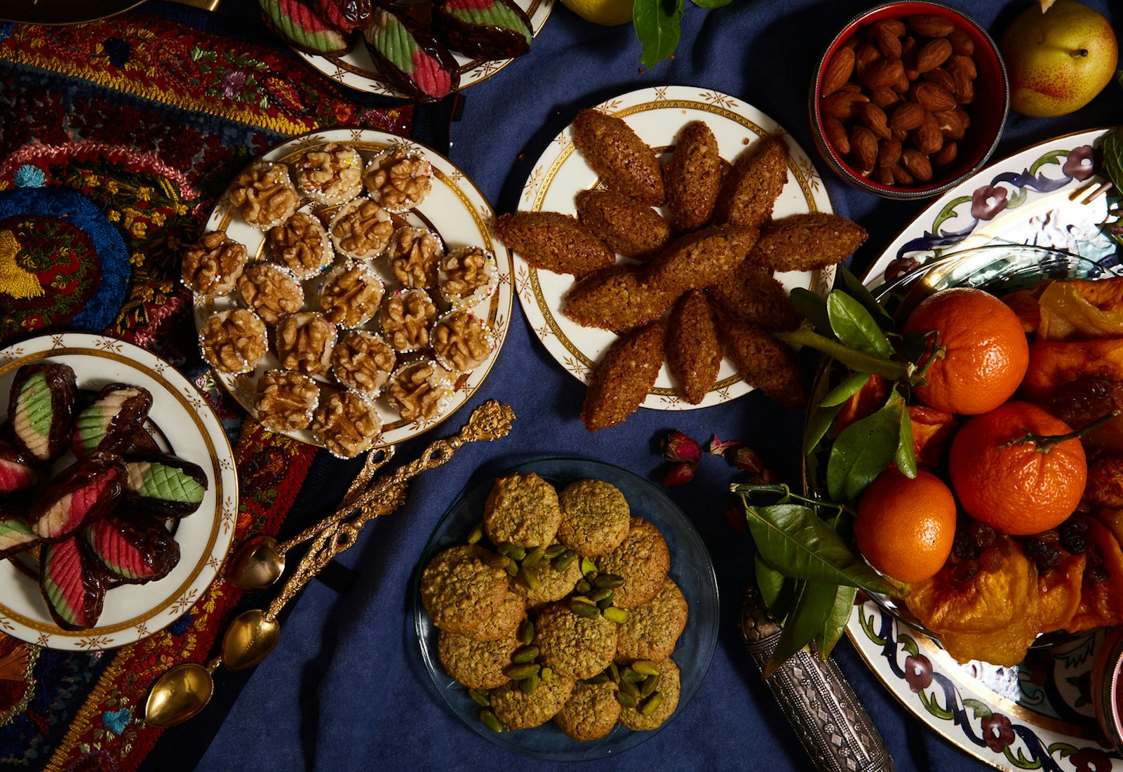Petits gateaux on blue plate surrounded by various cookies, nuts and fruits atop blue tablecloth.