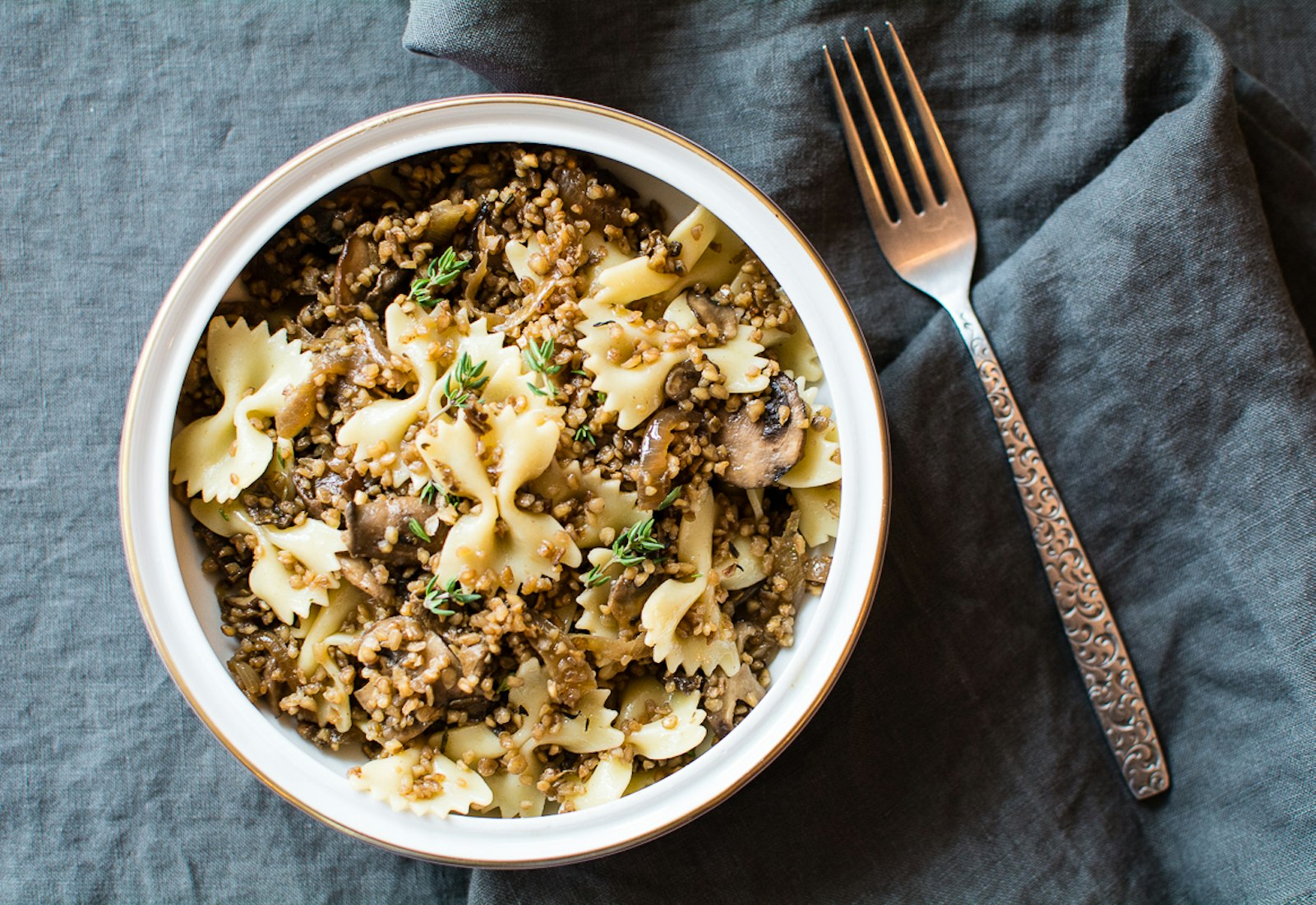 Kasha varnishes with mushrooms sprinkled with fresh thyme in white bowl atop grey cloth. 