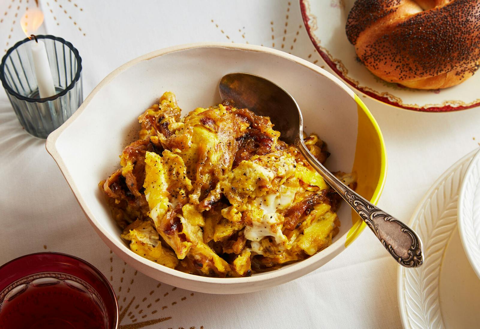 Tsibeles in cream-colored bowl alongside seeded challah, candle and red wine atop white tablecloth.