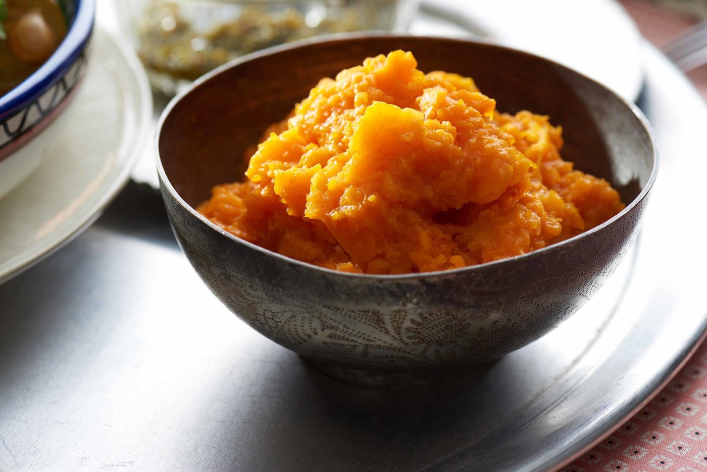 Mashed pumpkin in textured bowl on large tray, atop pink tablecloth.