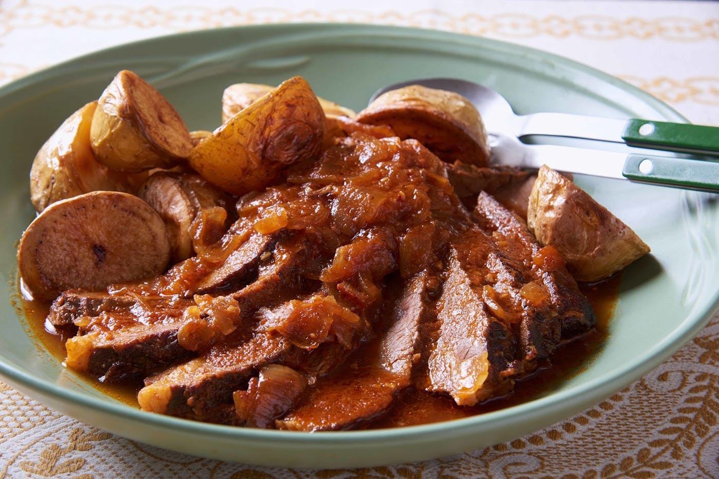 Sliced beef brisket coated with sauce alongside roasted baby potatoes in muted green dish with green serving utensils.