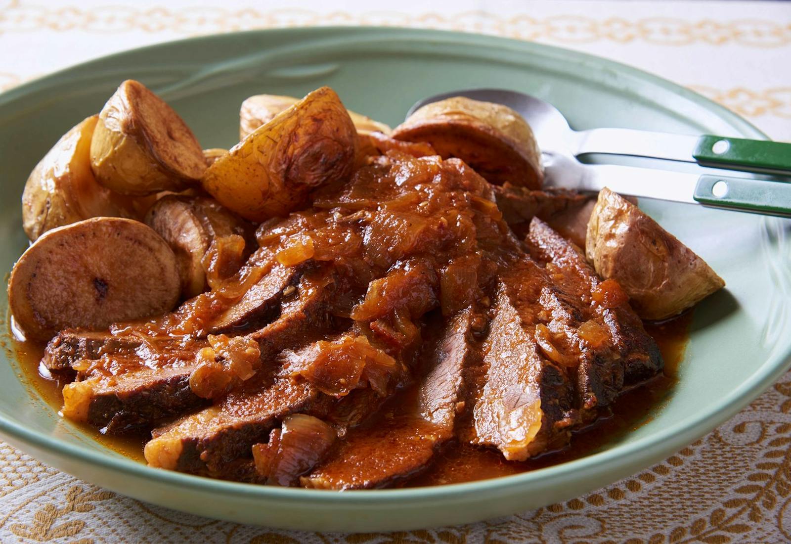 Sliced beef brisket coated with sauce alongside roasted baby potatoes in muted green dish with green serving utensils.