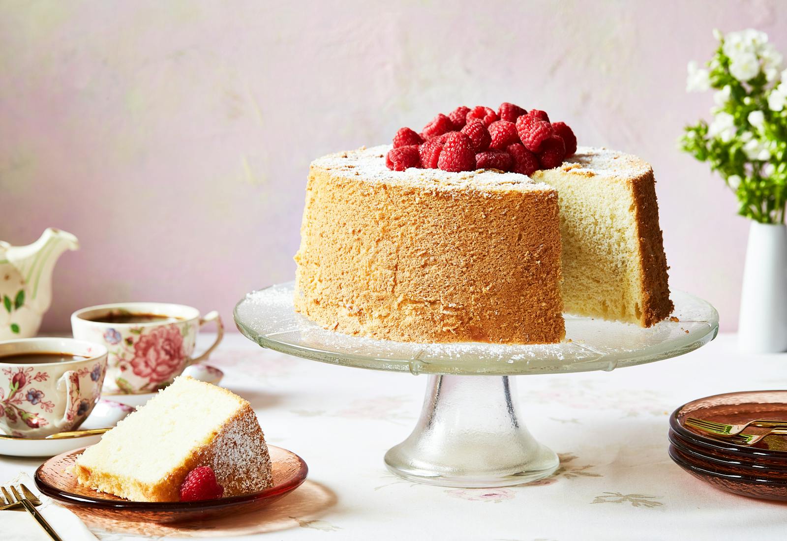 Chiffon cake with raspberries on top on a clear cake stand with a slice beside it.