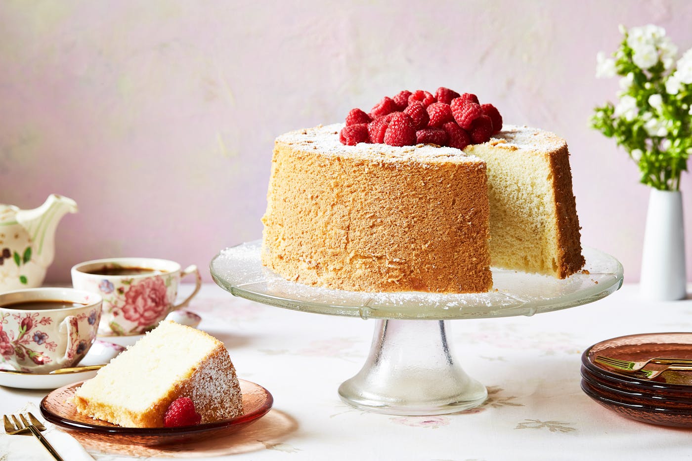 Chiffon cake with raspberries on top on a clear cake stand with a slice beside it.