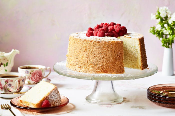 Chiffon cake with raspberries on top on a clear cake stand with a slice beside it.