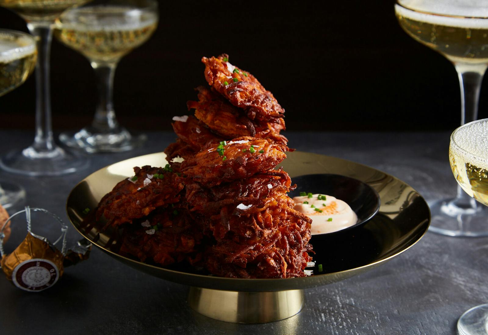 Stack of sweet potato latkes sprinkled with salt and chives alongside dish of sriracha creme fraiche, surrounded by sparkling white wine atop black table.