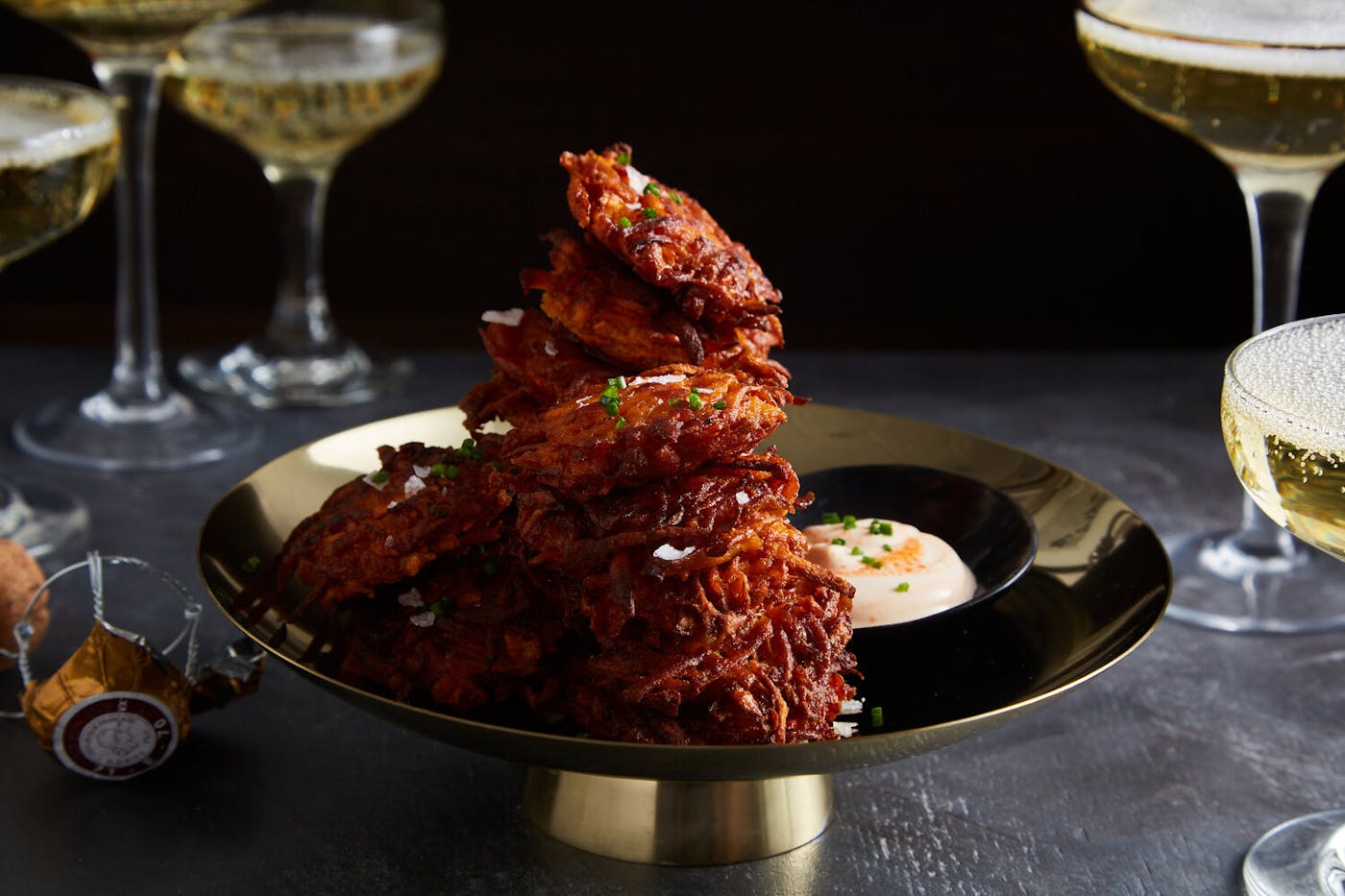 Stack of sweet potato latkes sprinkled with salt and chives alongside dish of sriracha creme fraiche, surrounded by sparkling white wine atop black table.