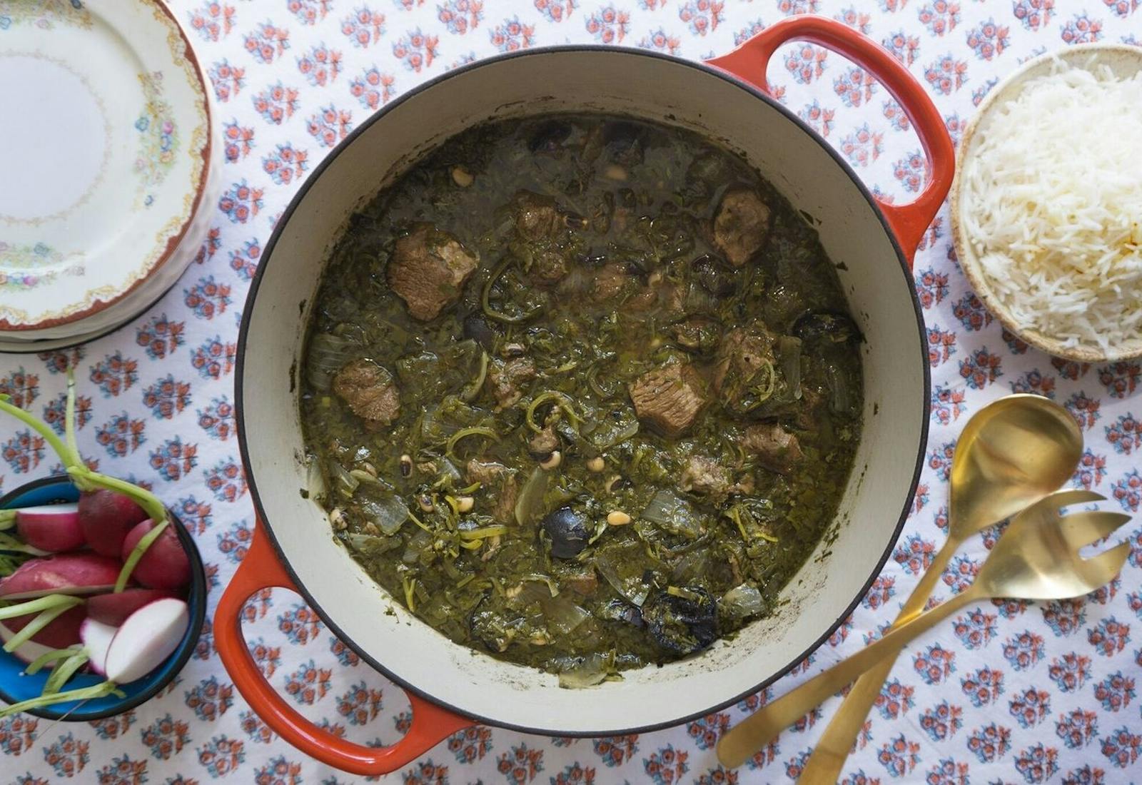 Ghormeh sabzi in white pot with red handles alongside gold serving utensils and bowls of white rice and fresh radishes, atop floral tablecloth.