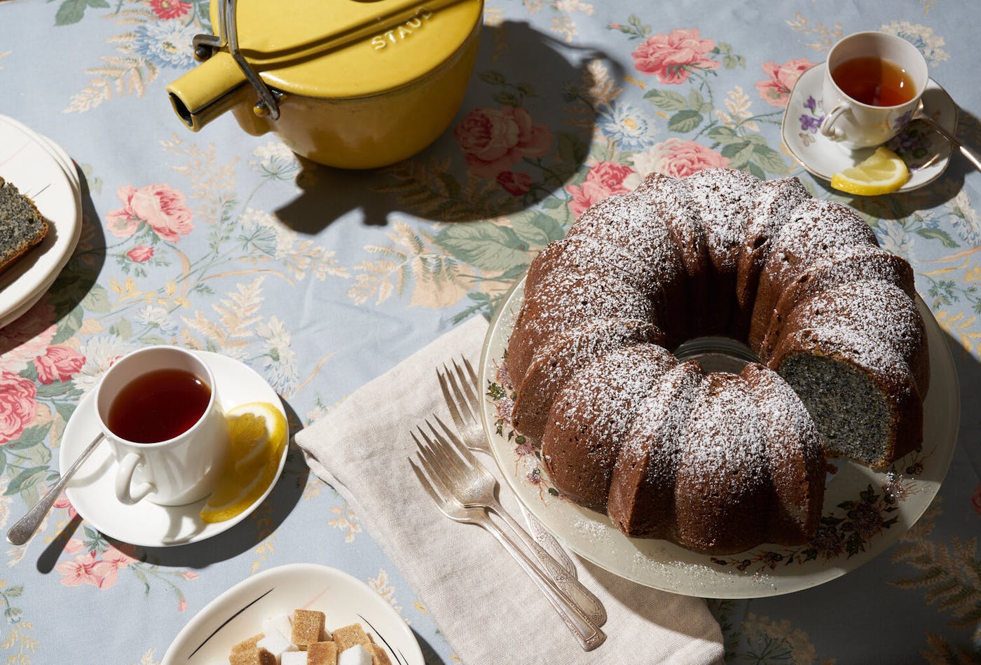 Poppy seed cake alongside tea with sugar cubes atop floral tablecloth.