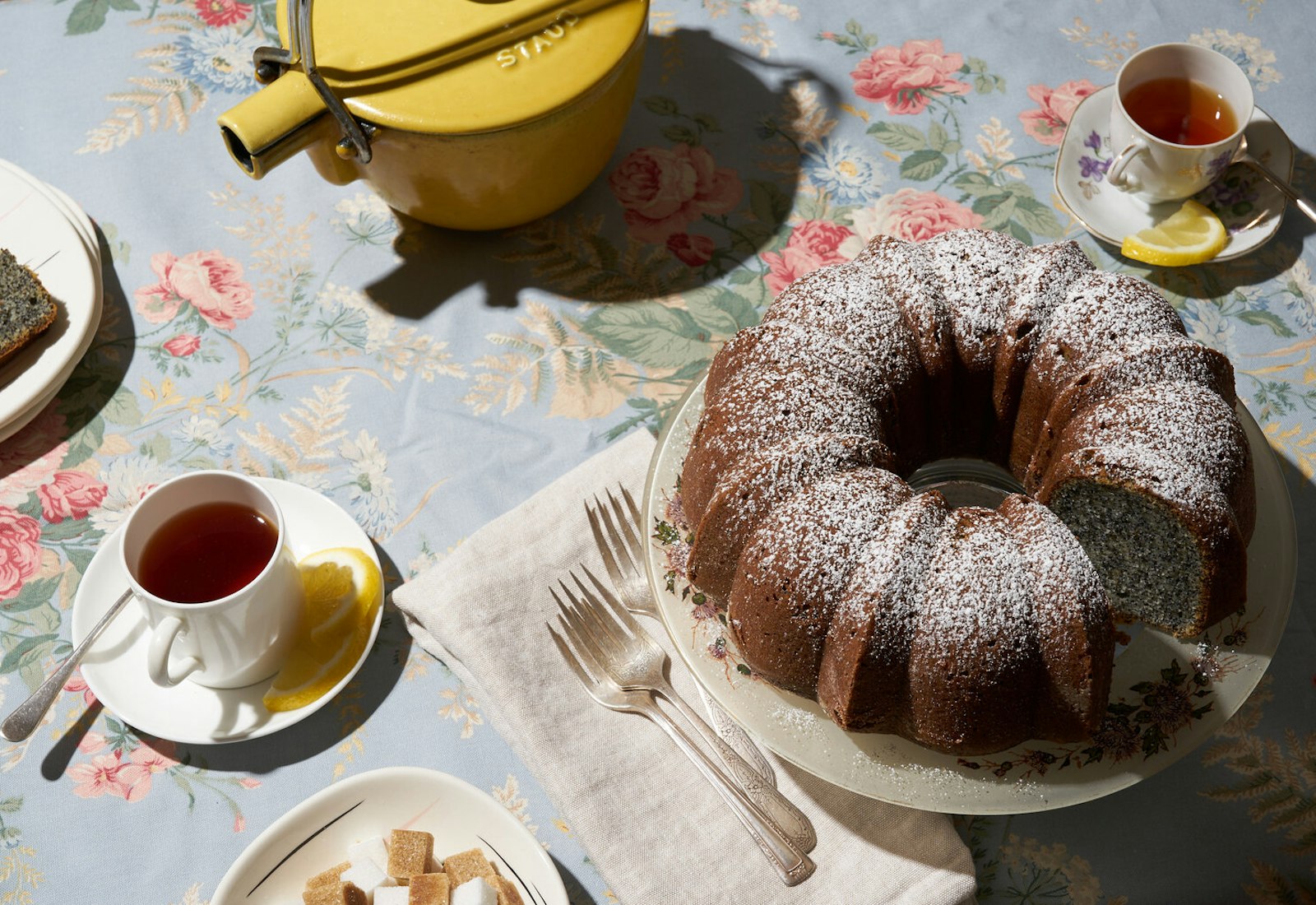 Poppy seed cake alongside tea with sugar cubes atop floral tablecloth.