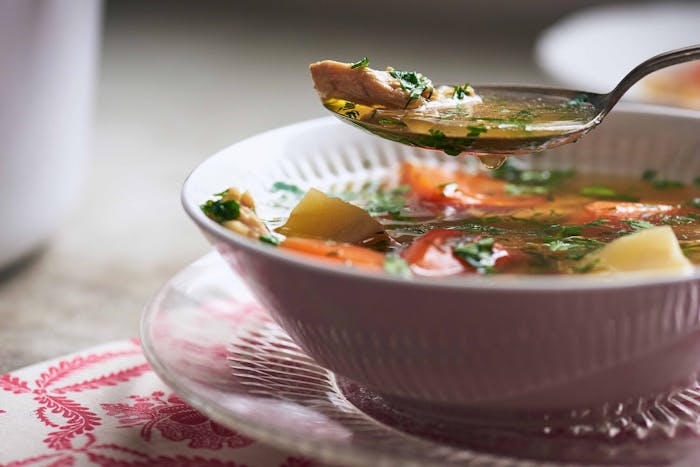 Chicken soup in white bowl with spoonful of broth coming out of bowl atop pink placemat.