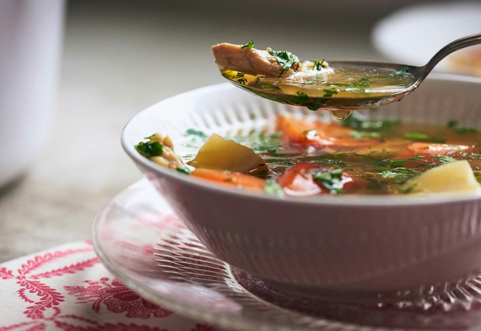 Chicken soup in white bowl with spoonful of broth coming out of bowl atop pink placemat.