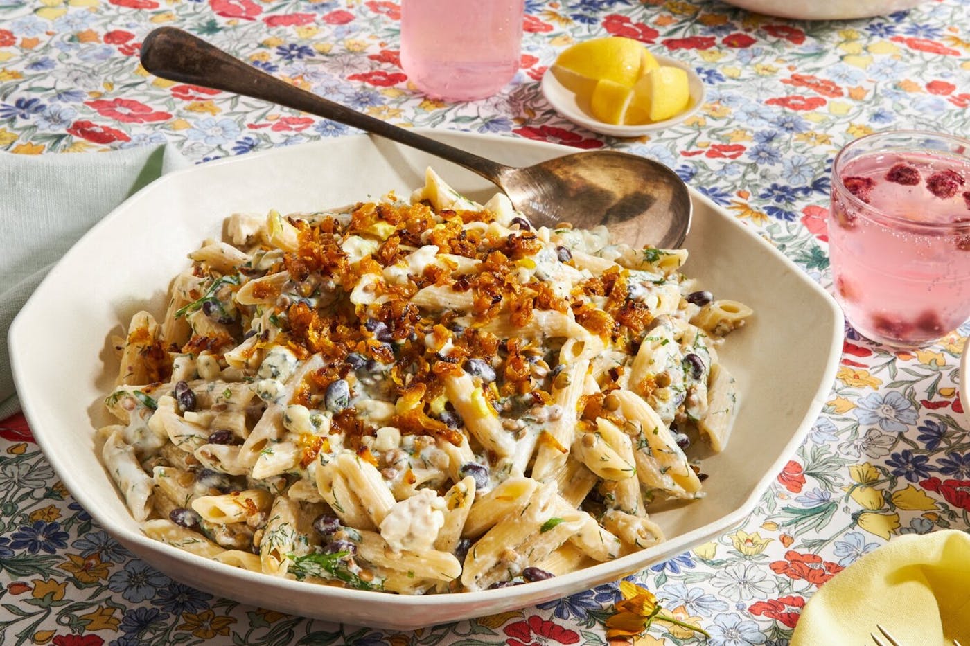 Pasta garnished with fried onions, bowl of lemon wedges and raspberry cocktails atop floral tablecloth. 