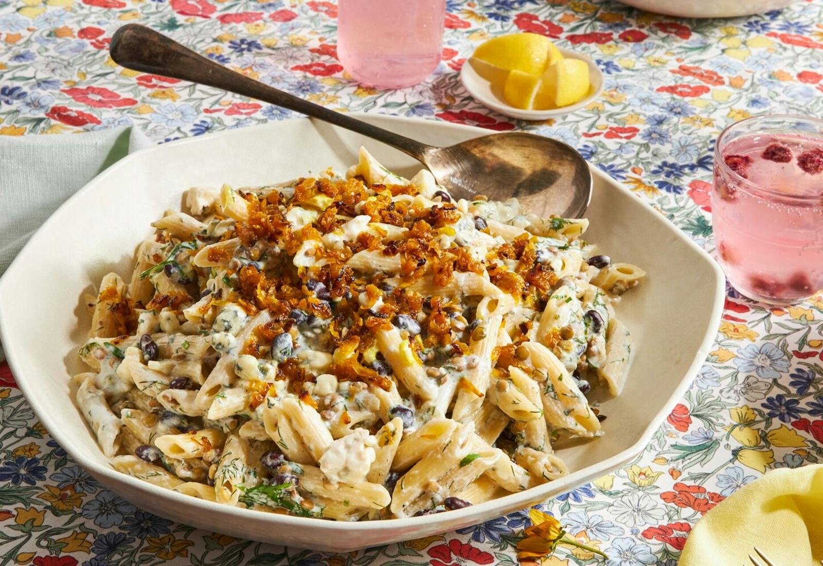 Pasta garnished with fried onions, bowl of lemon wedges and raspberry cocktails atop floral tablecloth. 