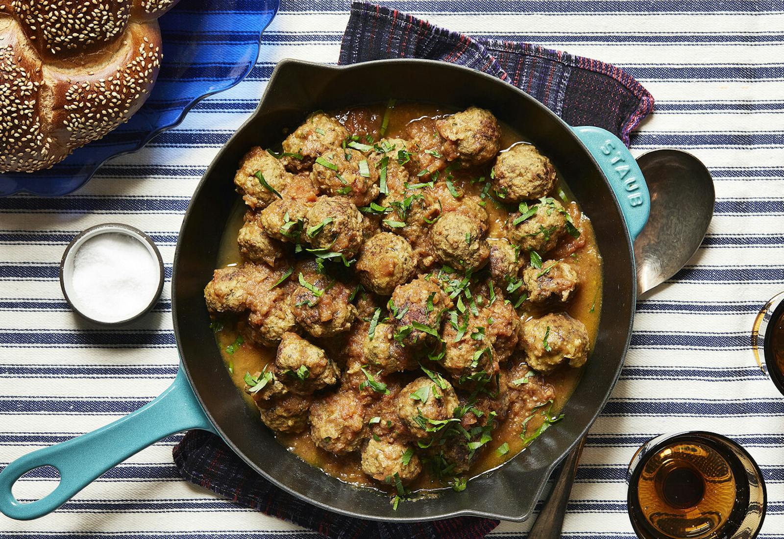 Meatballs with sliced parsley in cast iron skillet, seeded challah atop striped tablecloth.
